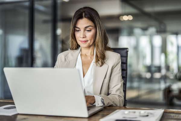 一位穿着浅色西装外套和衬衫的女士坐在木质桌前，专注地看着笔记本电脑
